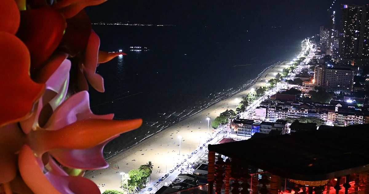 This is a stunning nighttime aerial view of Pattaya's coastline in Thailand. The photo shows the curved shoreline with its illuminated beachfront, where the golden sand beach is lit up against the dark ocean waters. The urban landscape feat