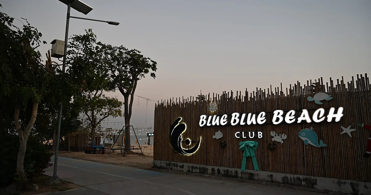 This photo shows the entrance to "Blue Blue Beach Club" in what appears to be Ban Chang, Rayong Province, Thailand. The sign is mounted on a rustic bamboo or wooden fence barrier that runs along the roadside. The sign features the name "BLU