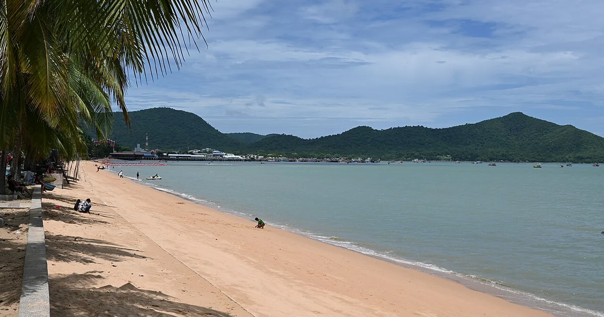 This photo shows a beautiful coastal scene with a long stretch of sandy beach alongside calm, grayish-blue waters. The beach has a light tan color and appears relatively uncrowded, with just a few visitors scattered along the shore. On the 