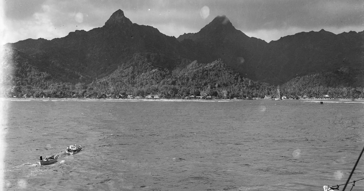 Taken from a ship. Two small boats are in the middleground on the left. In the background is a stretch of populated coastline with steep landscape rising up behind.
